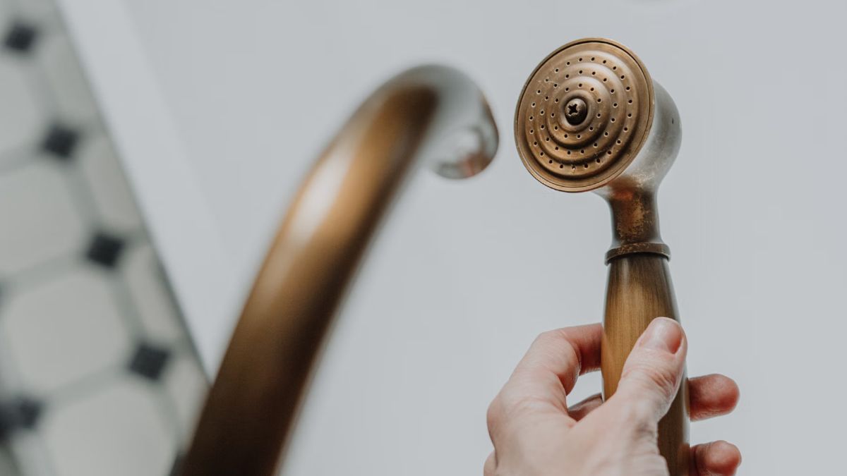 Hand holding a brass shower handset next to a brass bath spout