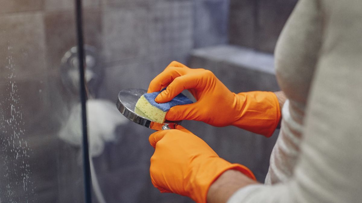 Hands in orange rubber gloves cleaning a shower head with a sponge