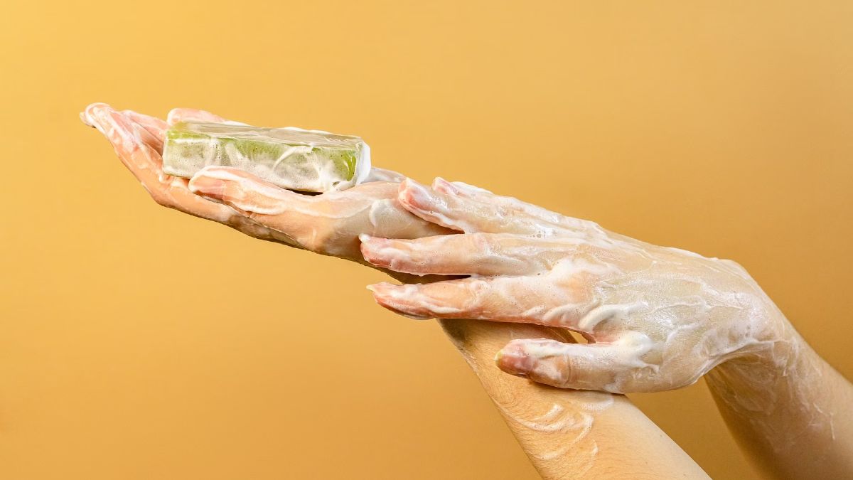 Hands covered in soap lather holding a clear bar of soap against a yellow background