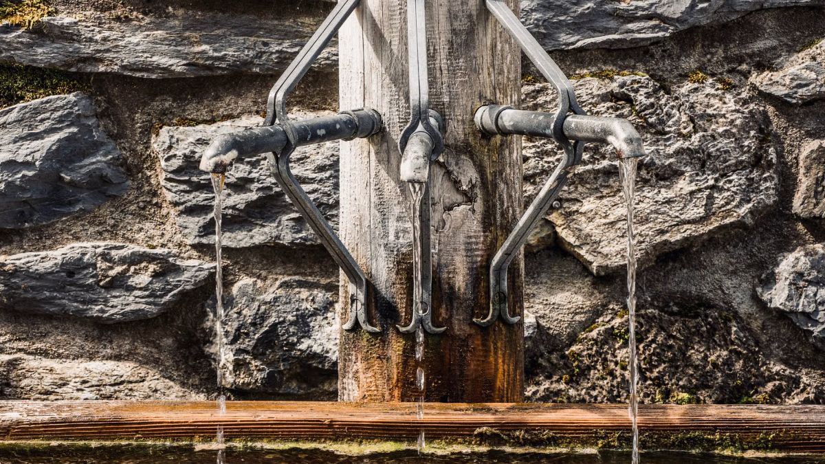 Two old metal taps on a wooden post running spring water in front of a stone wall